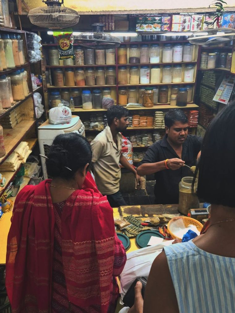 Customers at the counter of a spice shop in the market while a shopkeeper scoops spices from a jar, with shelves full of spice jars behind him