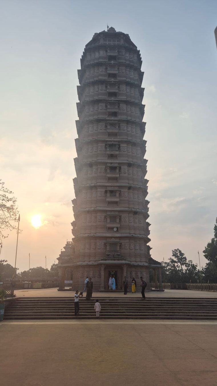 A tall, slender stone temple tower with many tiers, the low sun beside it and a few people on the steps in front