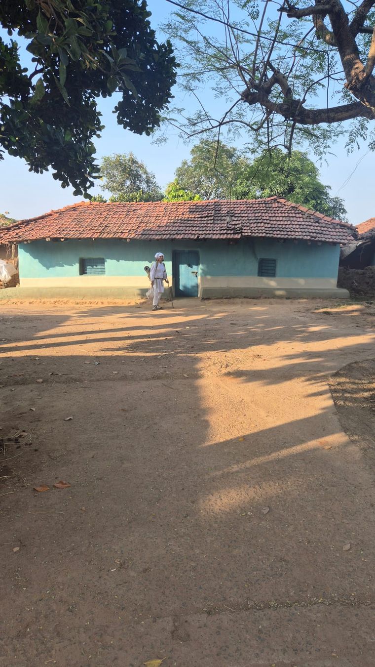 A low house with light blue walls, a blue door and a sloping roof of orange tiles under big trees; in front of it someone in white with a staff stands in a sandy yard full of shadows