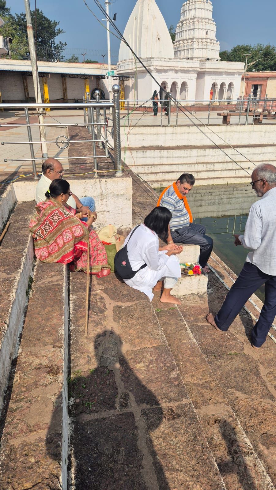 Yasha in white crouches on the stone steps of a temple tank with a small plate of flowers in front of her, next to Diederik in an orange scarf, an older man and woman and a man with glasses; white temples in the background