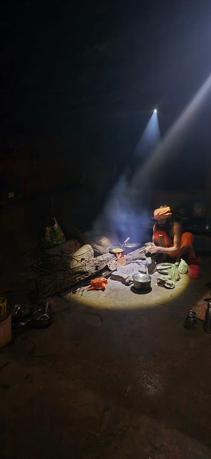 A man in orange clothes and an orange turban crouches in the dark by a chulha making roti, lit by the beam of a torch with smoke rising above him