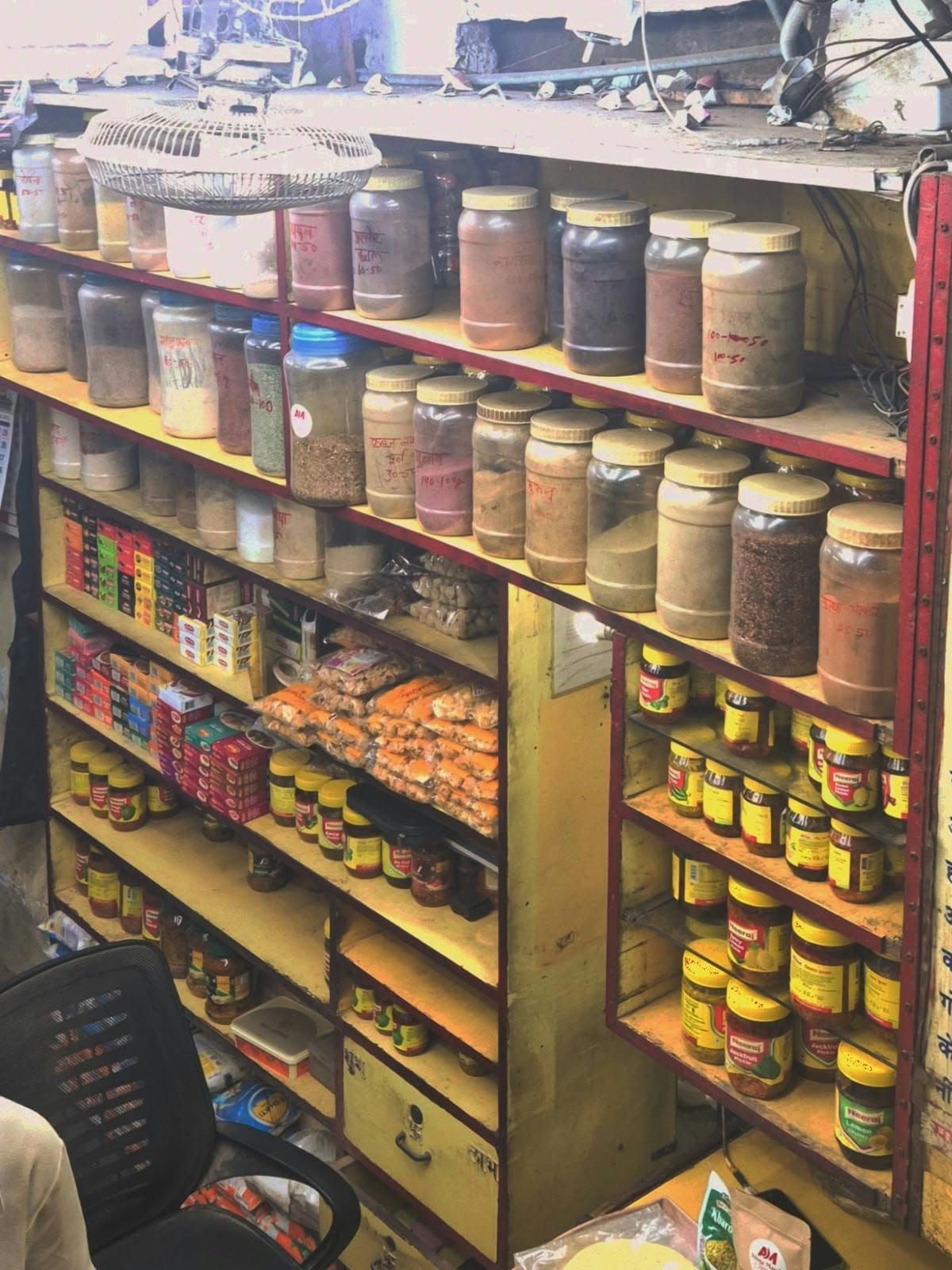 Yellow shelves in a small shop full of spice jars with prices written on them, and rows of pickle jars below