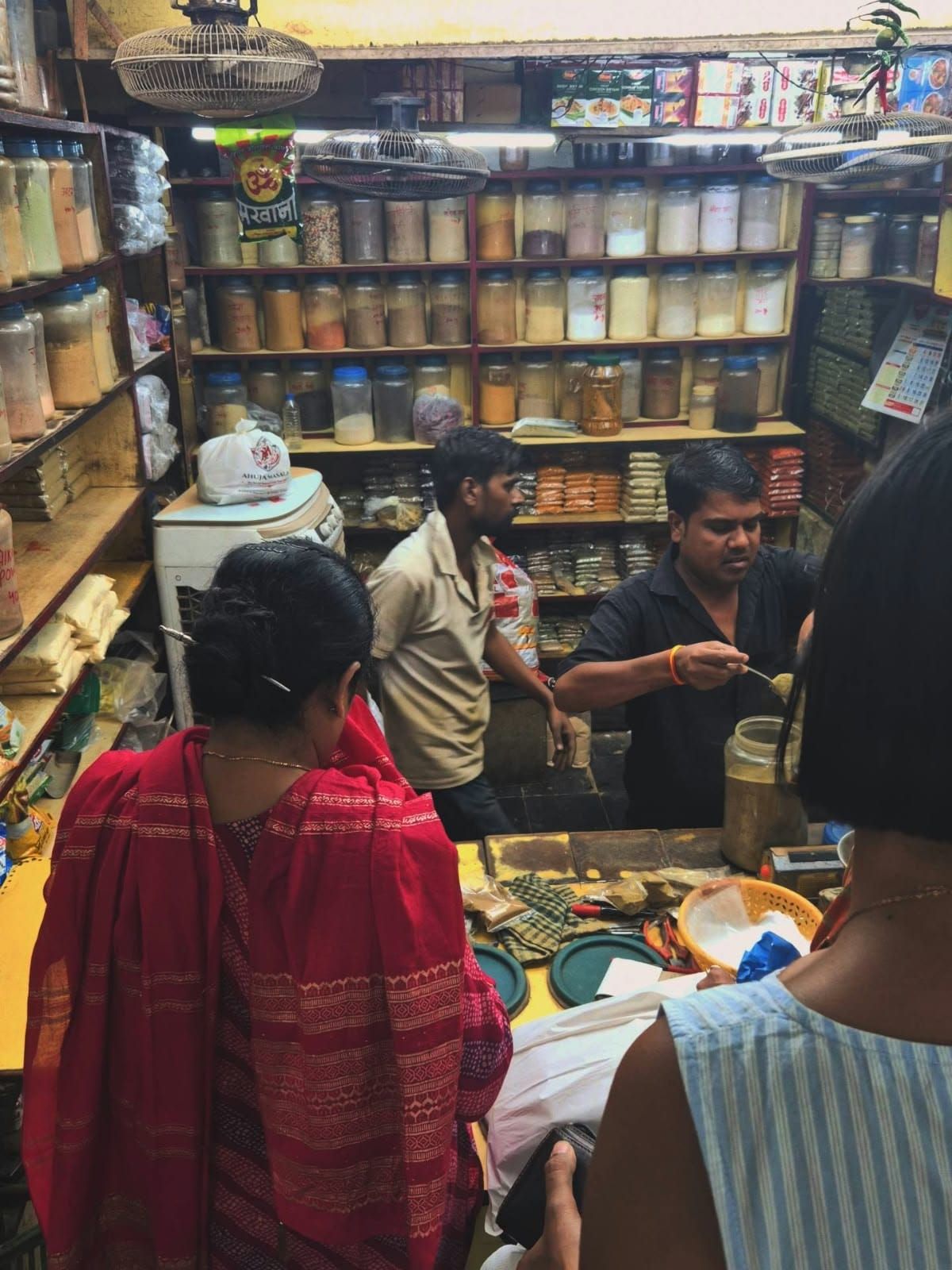 Customers at the counter of a spice shop in the market while a shopkeeper scoops spices from a jar, with shelves full of spice jars behind him