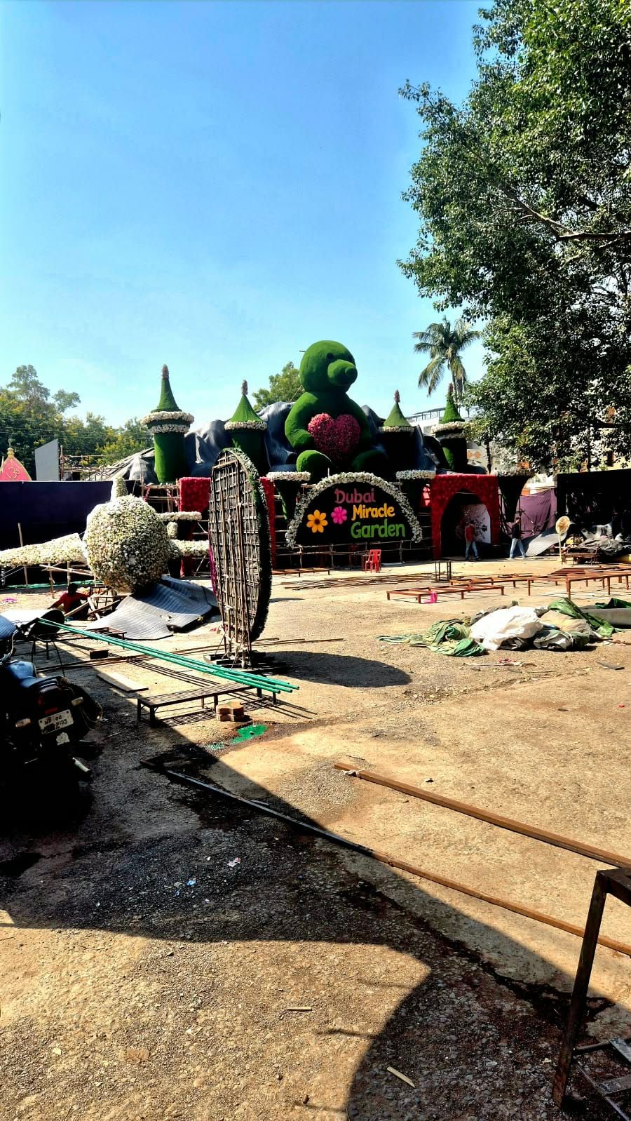 A large flower structure being built on an open lot: a giant green teddy bear with a pink heart, green turrets and an arch reading Dubai Miracle Garden, with building materials on the ground in front