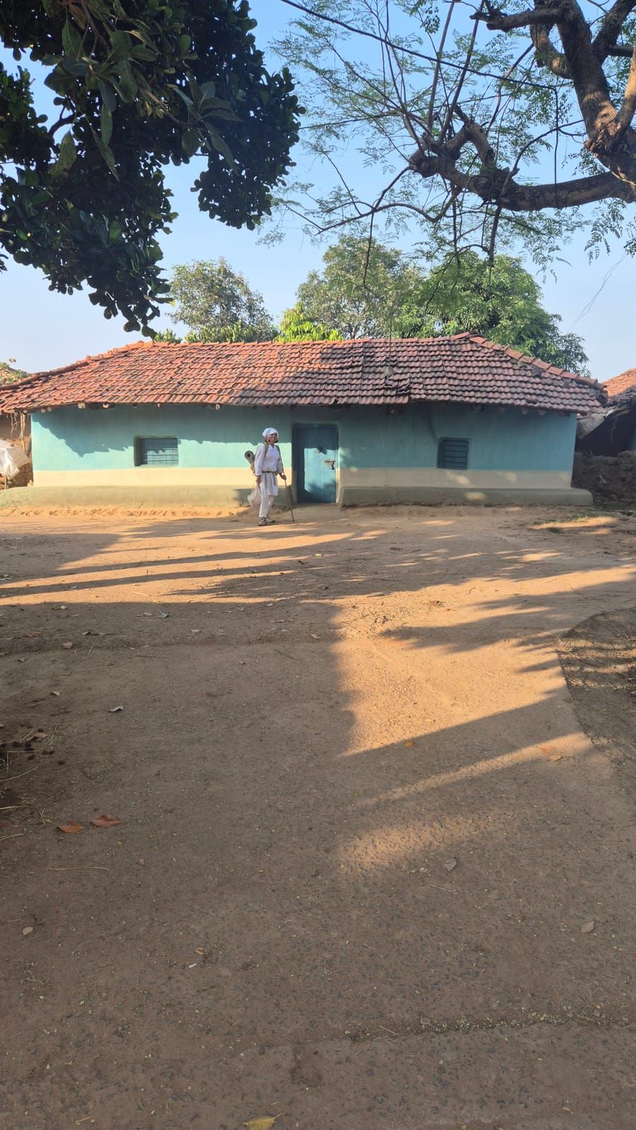 A low house with light blue walls, a blue door and a sloping roof of orange tiles under big trees; in front of it someone in white with a staff stands in a sandy yard full of shadows