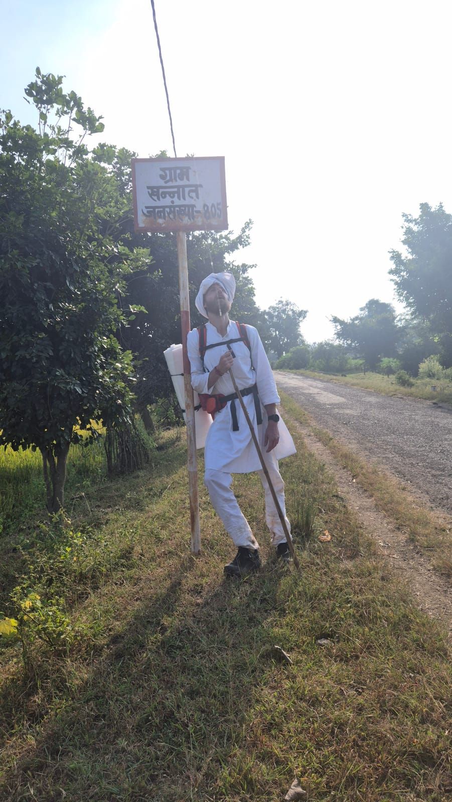 Diederik in white, with a white turban, a backpack and a long staff, leans against the post of a village sign in Hindi next to a country lane