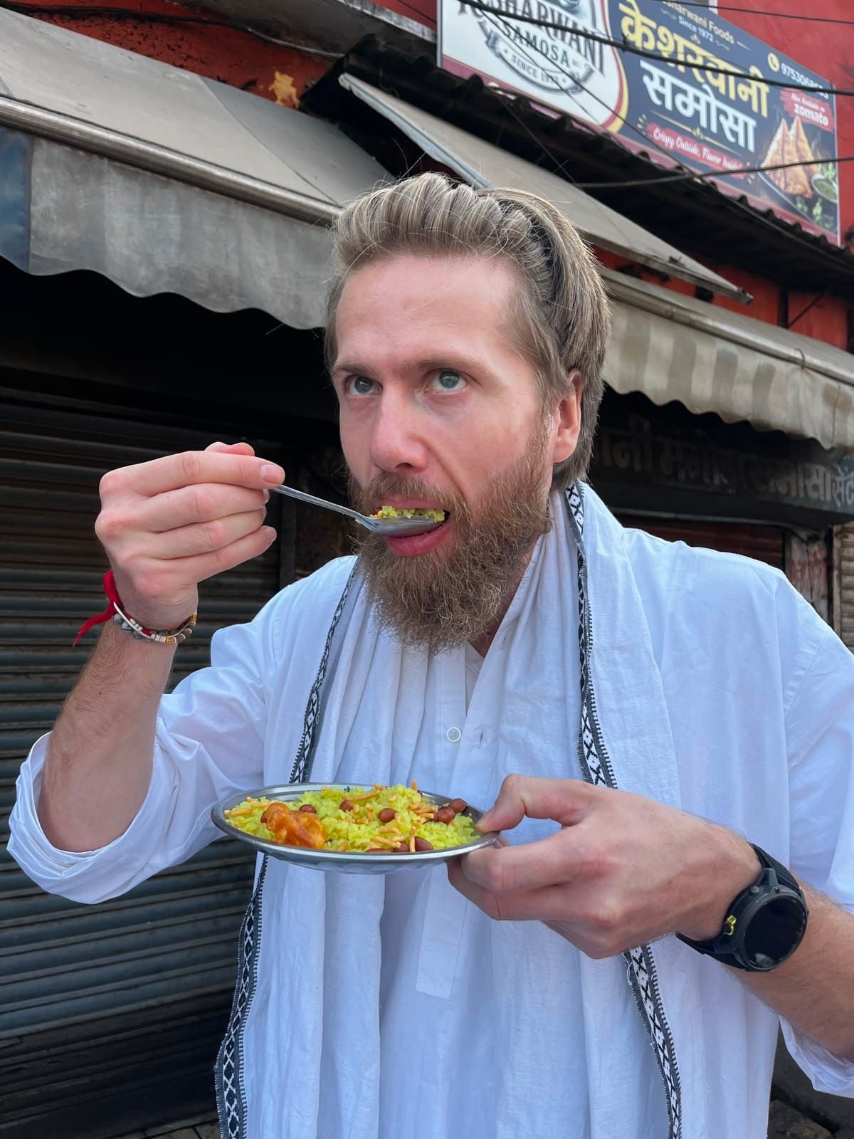 Diederik in a white kurta and scarf takes a bite of poha from a metal plate, in front of a closed shop with a samosa sign