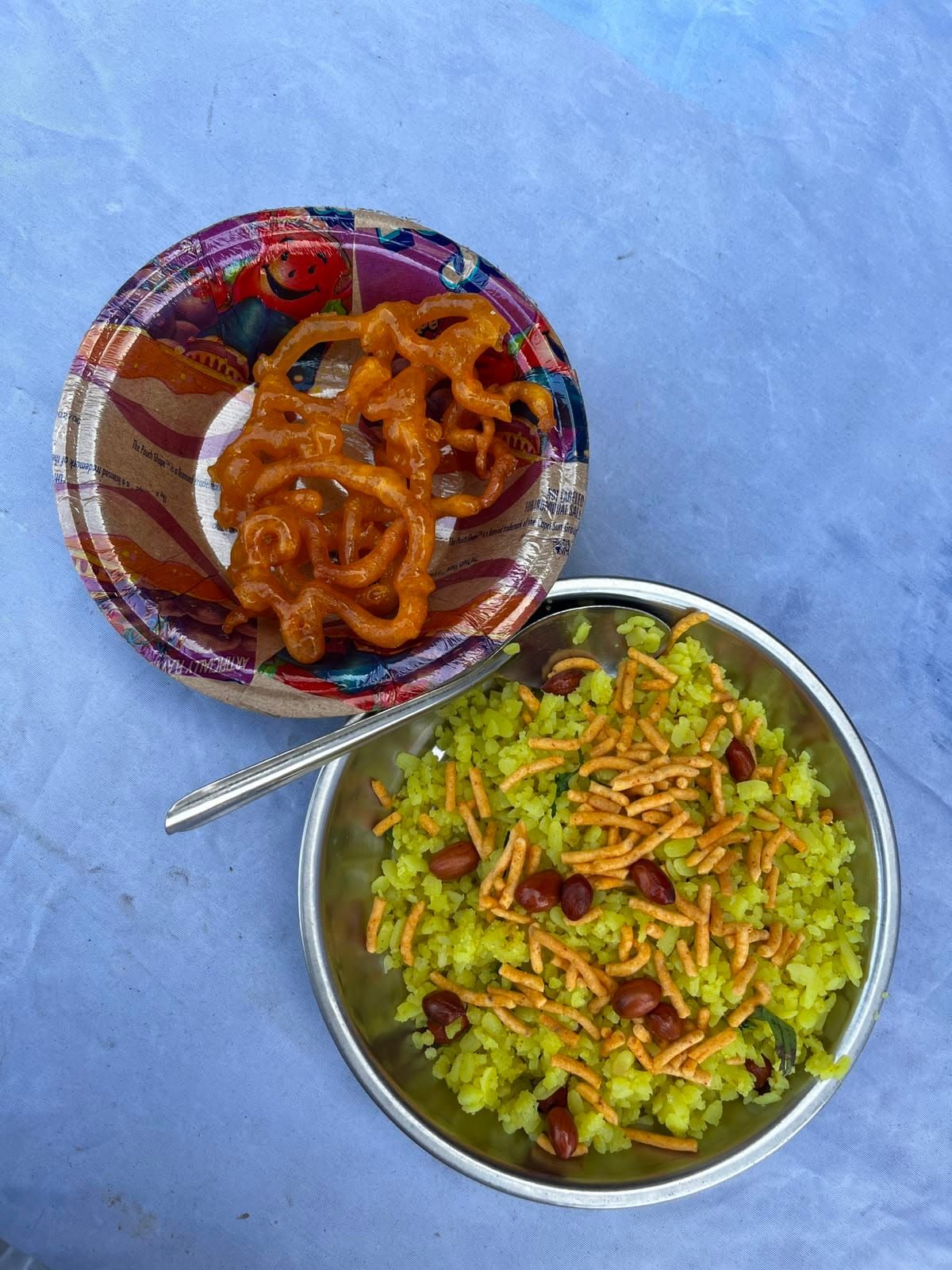 From above: a paper plate of orange jalebi and a metal plate of yellow poha with peanuts and sev