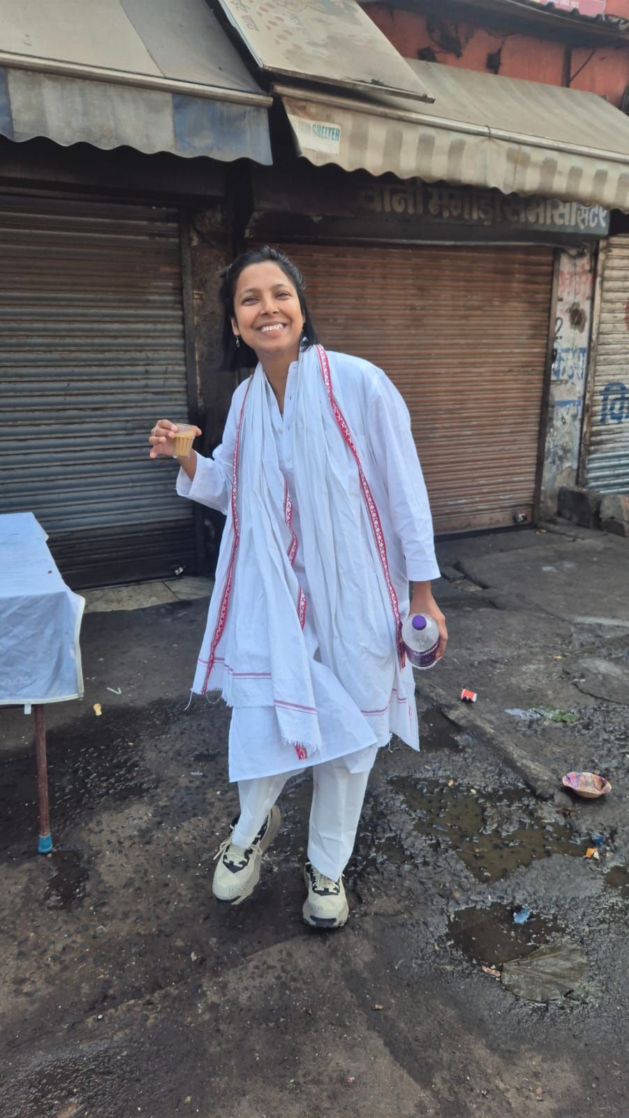 Yasha, dressed in white, laughs with a small glass of chai in one hand and a water bottle in the other, in front of closed shutters