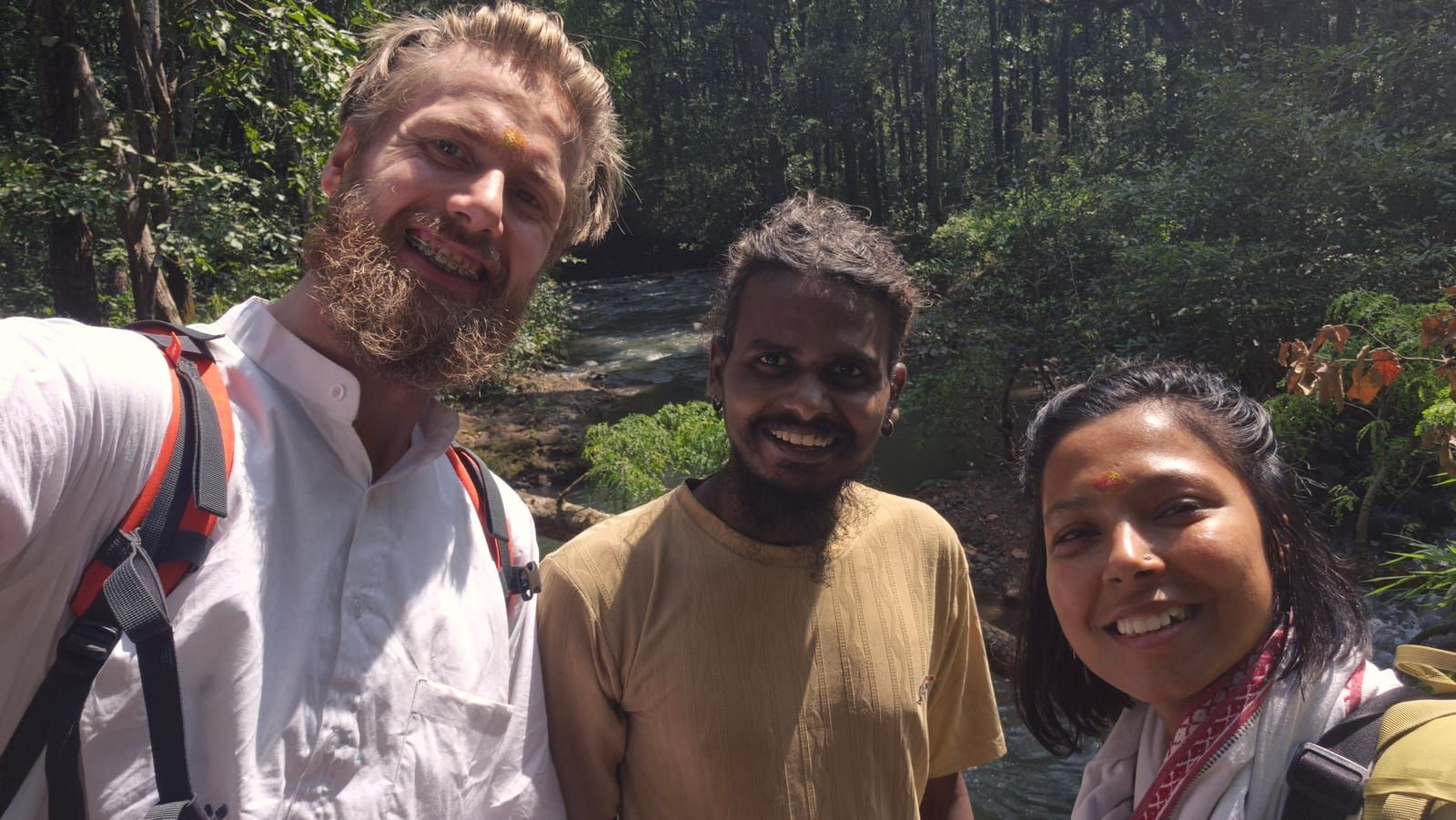 Selfie of Diederik with a beard and a white kurta, a smiling man with a beard in an olive T-shirt, and Yasha with a red dot on her forehead, by a flowing stream in the jungle
