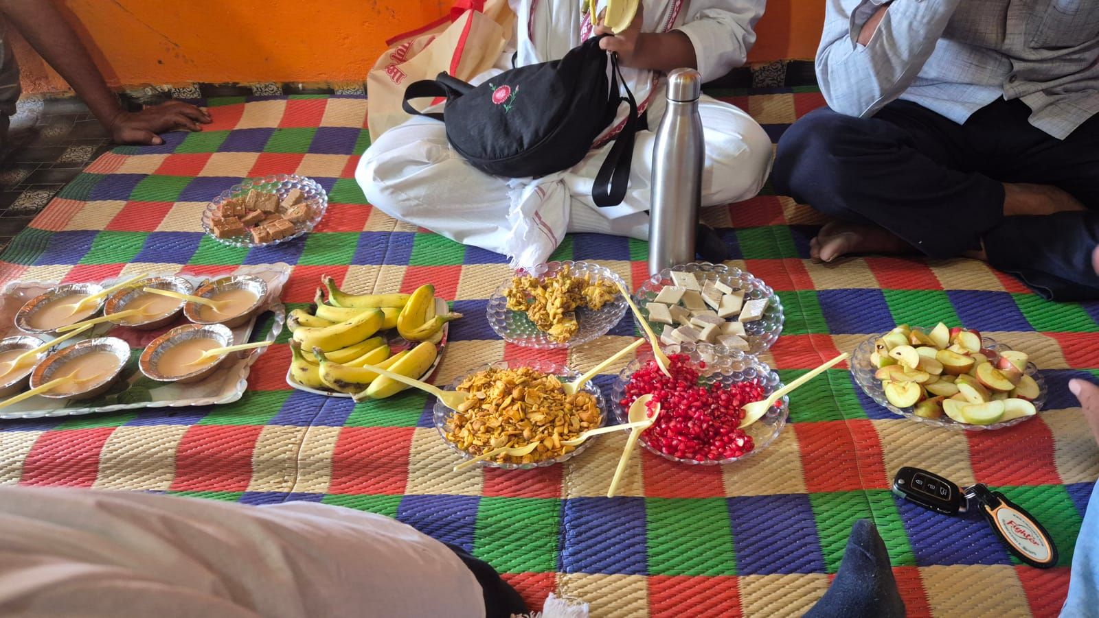 On a colourful checked mat on the floor: little bowls of chai, bananas, fried snacks, barfi, pomegranate seeds and apple slices