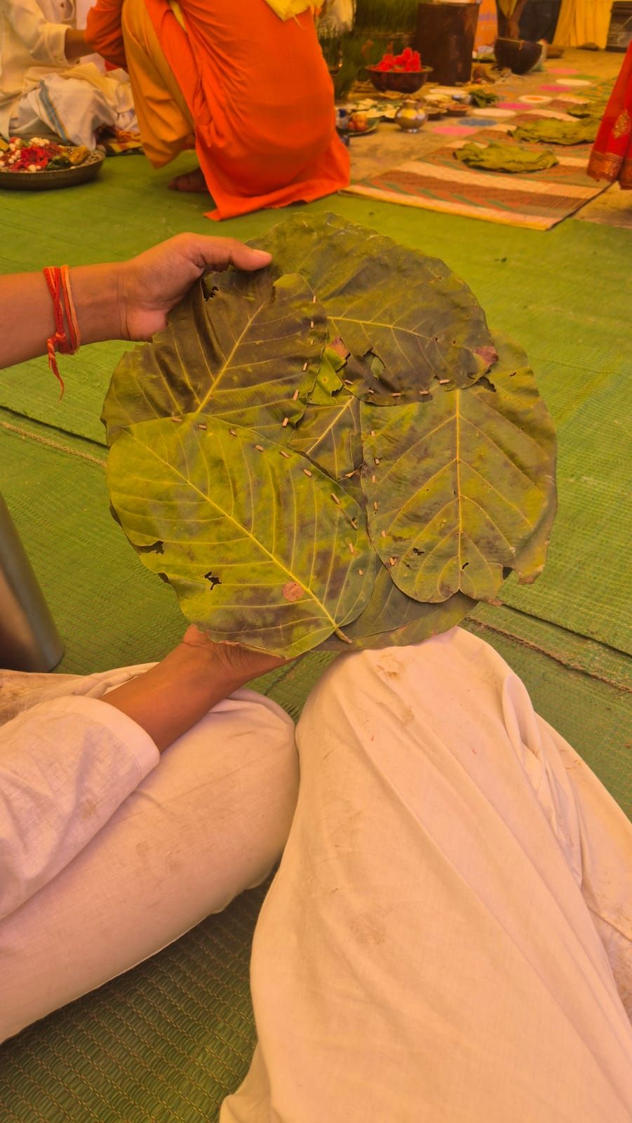 Someone in white holds up a round plate made of large green leaves pinned together with little wooden sticks; in the background there are bowls of flowers and offerings on the mats