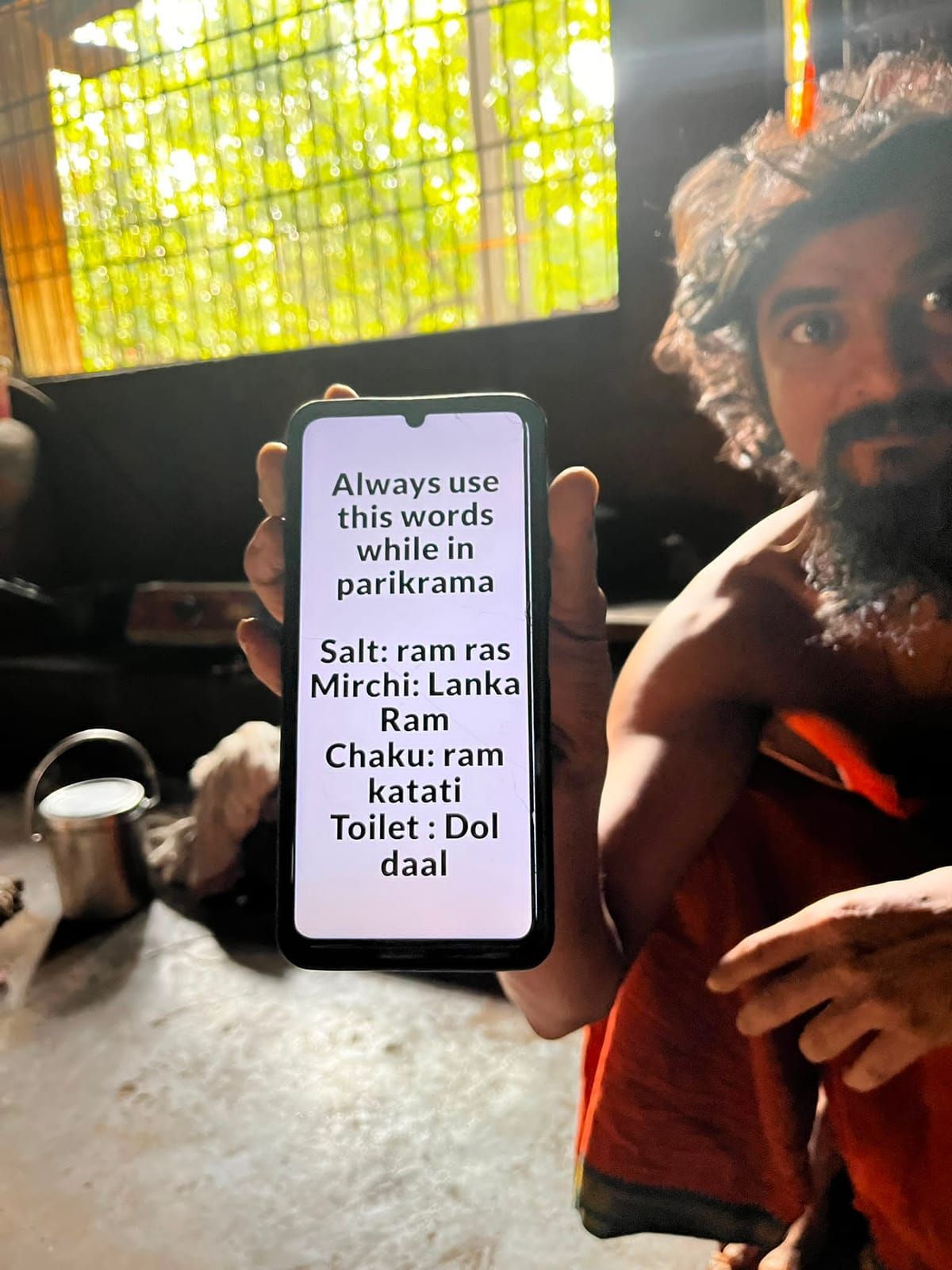 Vijay, with long curly hair and a beard, holds up his phone in the dim ashram with the text: Always use this words while in parikrama. Salt: ram ras, Mirchi: Lanka Ram, Chaku: ram katati, Toilet: Dol daal