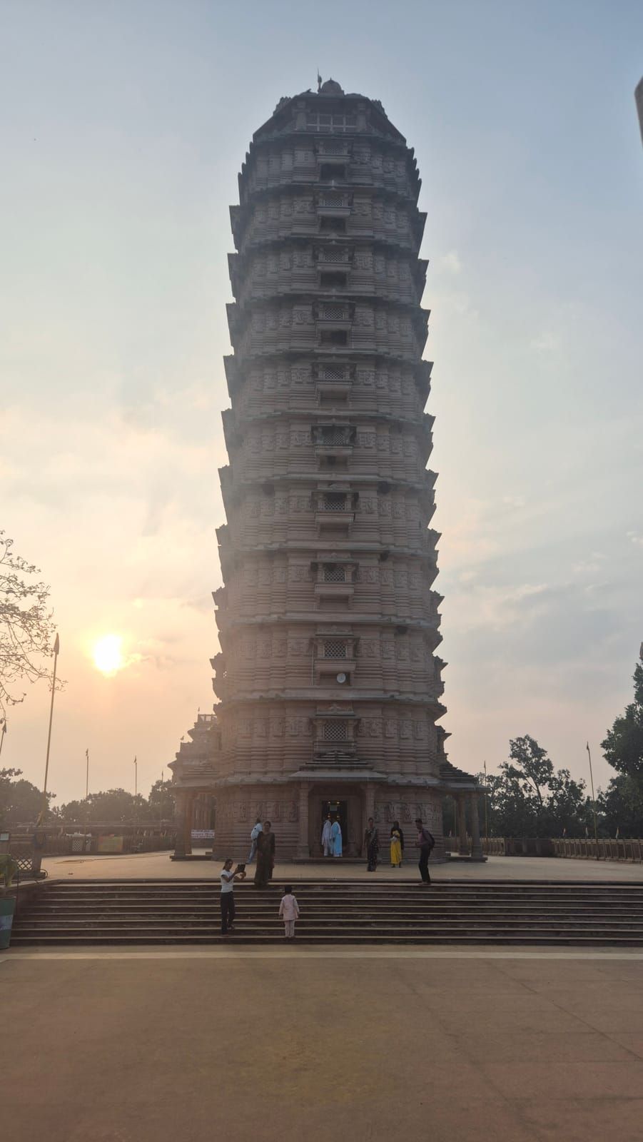 A tall, slender stone temple tower with many tiers, the low sun beside it and a few people on the steps in front