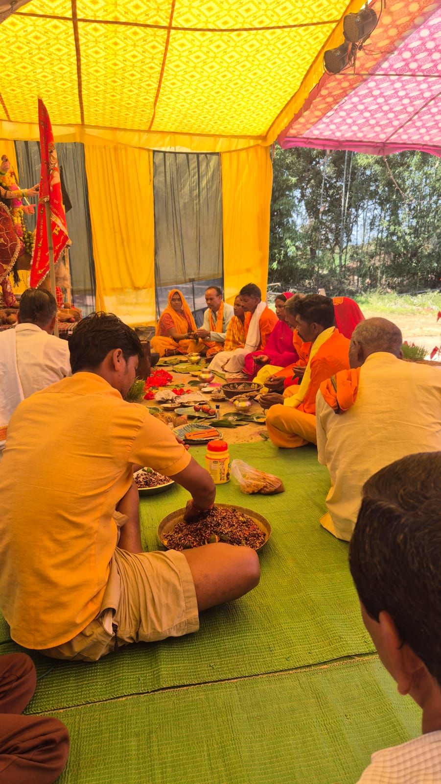 Under a yellow tent, people in orange, yellow and red clothes sit on green mats in front of a decorated statue of Durga; in the foreground a man in a yellow shirt mixes herbs in a large bowl